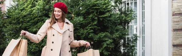 Joyeuse femme en béret rouge et trench coat beige tenant des sacs à provisions à l'extérieur, bannière — Photo de stock