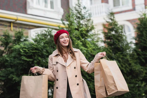 Young smiling woman in red beret and beige trench coat holding shopping bags outside — Stock Photo