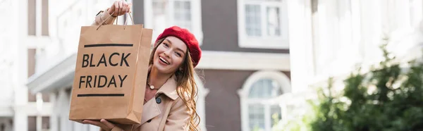 Cheerful young woman in red beret and trench coat holding shopping bag with black friday lettering outside, banner — Stock Photo