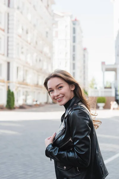 Mujer joven con estilo en cuello alto negro y chaqueta de cuero sonriendo en la calle urbana de Europa - foto de stock