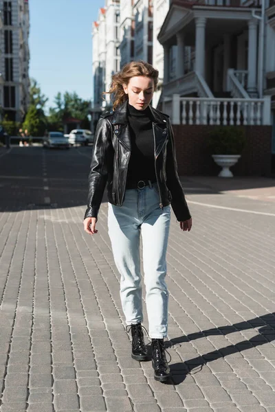 Comprimento total de elegante jovem mulher em gola alta preta, botas e jaqueta de couro andando na rua urbana da Europa — Fotografia de Stock
