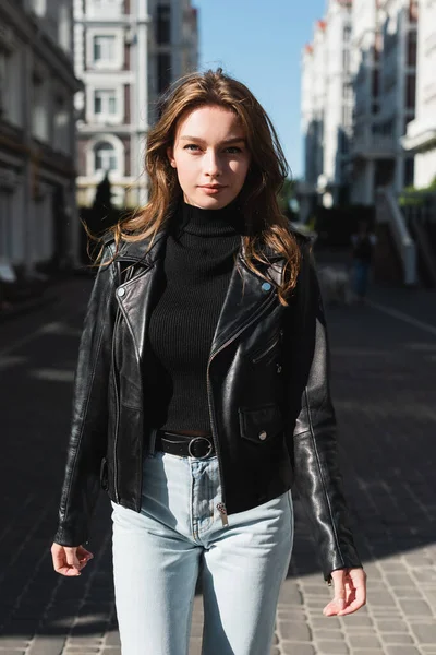 Mujer joven en cuello alto negro y chaqueta de cuero mirando a la cámara en la calle urbana de Europa - foto de stock