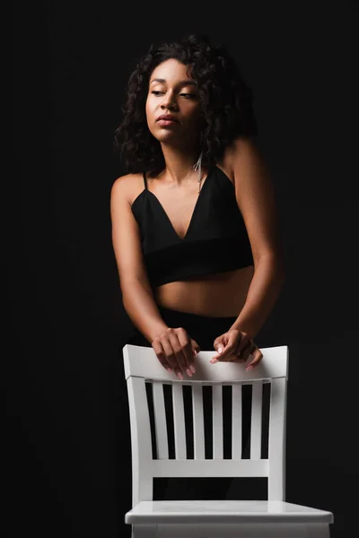 African american woman in sexy satin crop top posing near chair isolated on black — Stock Photo