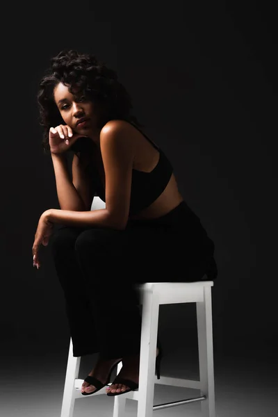 Sexy african american woman in satin crop top sitting on white chair on black — Stock Photo