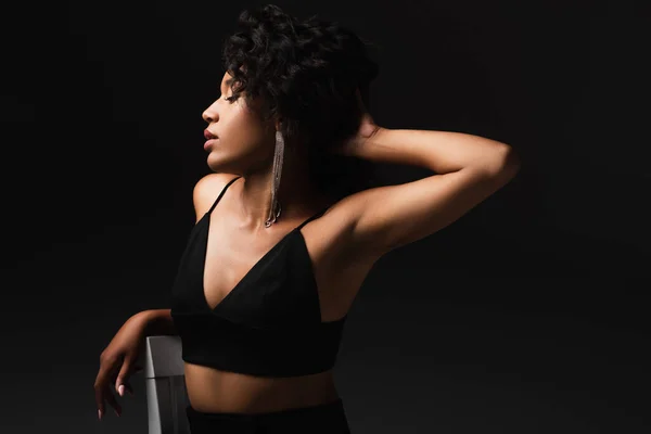 Seductive young african american woman in satin crop top posing near chair isolated on black — Stock Photo