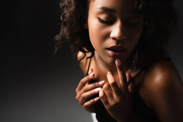 Seductive young african american woman in sexy crop top touching lip while posing on black — Stock Photo