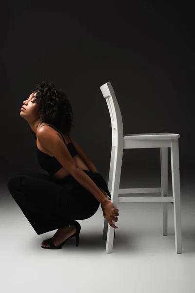 Side view of sexy african american woman in satin crop top and pants sitting near white chair while posing on black — Stock Photo