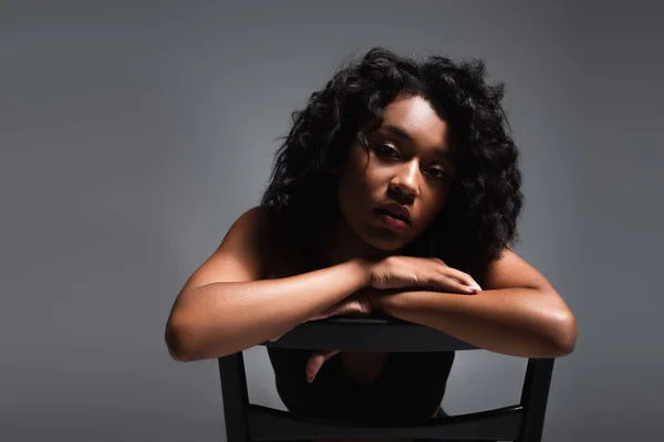 Young african american woman leaning on wooden chair while posing on grey — Stock Photo