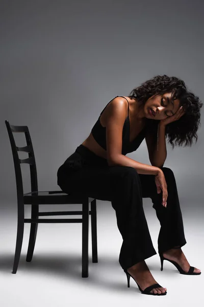 Full length of brunette african american woman in stylish outfit sitting on chair on grey — Stock Photo