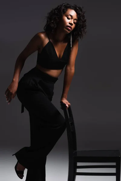 Curly african american woman in trendy outfit posing on wooden chair on dark grey — Stock Photo