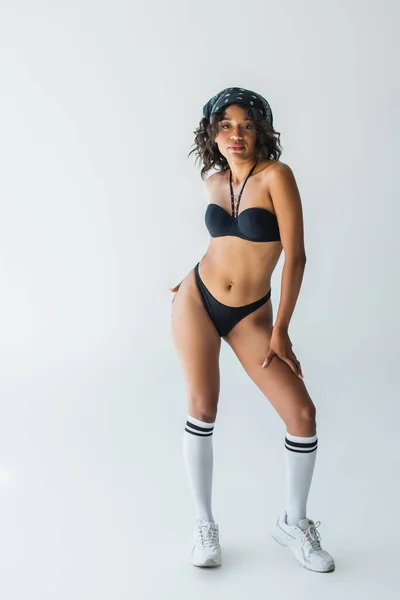 Pleine longueur de femme afro-américaine à la mode en maillot de bain, chaussettes hautes genou et baskets posant sur gris — Photo de stock