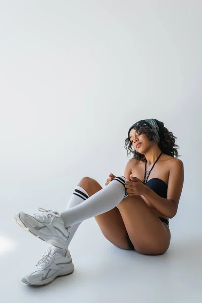 Longueur totale de femme afro-américaine élégante en maillot de bain et baskets ajustant le genou haut chaussette sur gris — Photo de stock