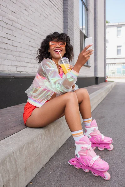 Full length of smiling african american woman in sunglasses and roller skates drinking orange juice while holding cellphone — Stock Photo