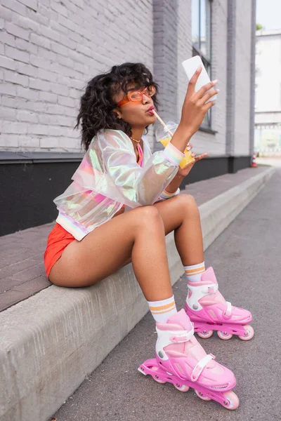 Longitud completa de la joven afroamericana en patines bebiendo jugo de naranja y tomando selfie - foto de stock