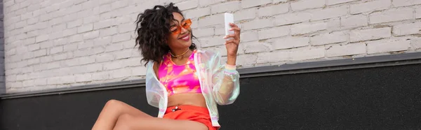 Femme afro-américaine souriante dans des lunettes de soleil en utilisant un smartphone à l'extérieur, bannière — Photo de stock