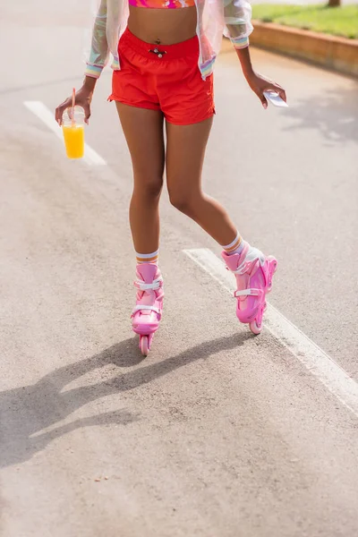 Vista recortada de la mujer afroamericana patinando en patines de ruedas mientras sostiene la taza de plástico y el teléfono celular - foto de stock