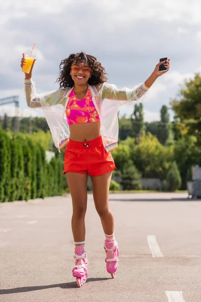 Longitud completa de alegre mujer afroamericana patinando en patines de ruedas mientras sostiene la taza de plástico y el teléfono inteligente con pantalla en blanco - foto de stock