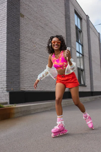 Mulher americana africana alegre em fones de ouvido sem fio patinação em patins de rolo enquanto segurando smartphone — Fotografia de Stock