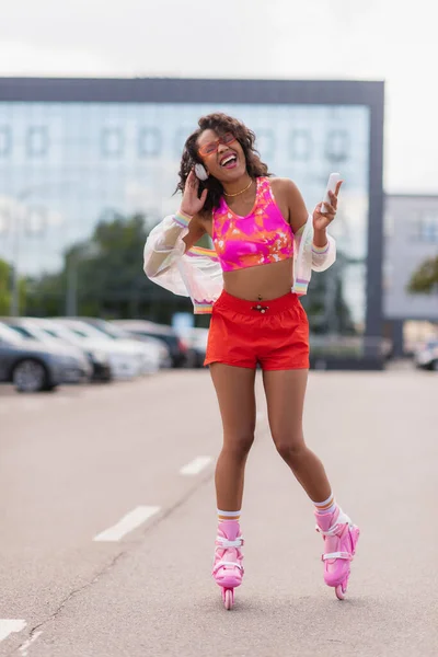 Femme afro-américaine joyeuse dans les écouteurs sans fil et sur les patins à roulettes chantant tout en tenant smartphone — Photo de stock