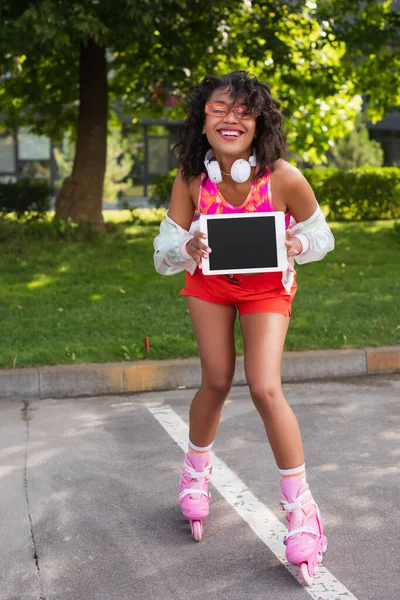 Mulher americana africana encaracolado em fones de ouvido sem fio e em patins sorrindo enquanto segurando tablet digital com tela em branco — Fotografia de Stock