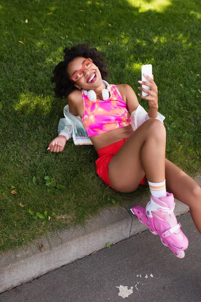 Mulher americana africana alegre em patins sorrindo enquanto segurando smartphone e descansando na grama — Fotografia de Stock