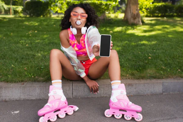 Blurred african american woman on roller skates blowing bubble gum while sitting on grass with smartphone — Stock Photo