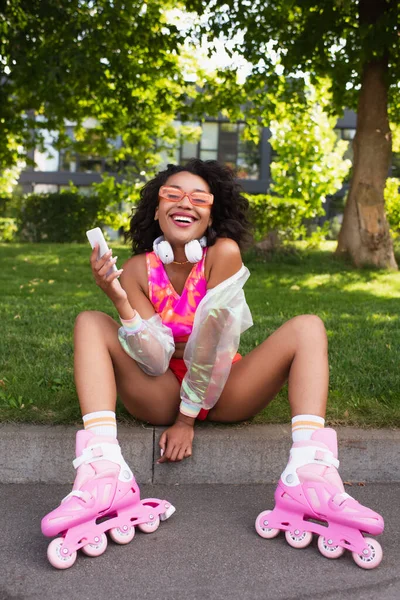 Счастливая африканская американка в наушниках и на роликовых коньках улыбается, держа в руках смартфон — стоковое фото