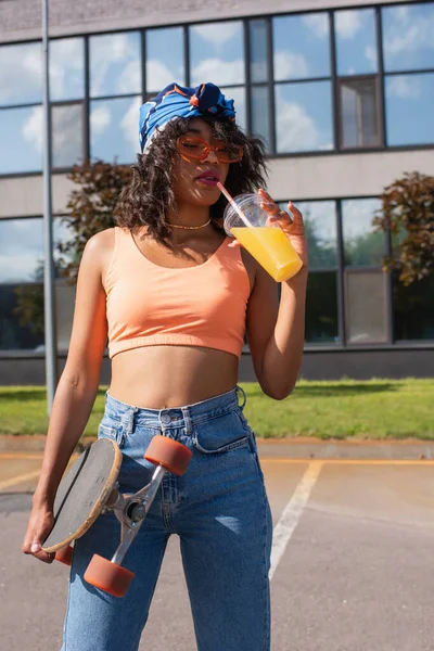 Young african american woman in headscarf drinking orange juice and holding longboard outside — Stock Photo