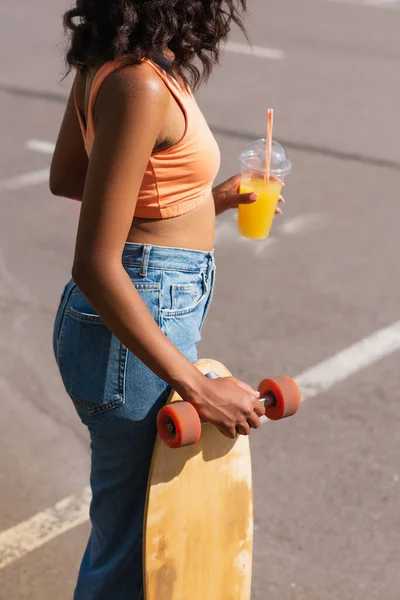 Vista cortada da mulher americana africana encaracolado segurando copo de plástico com suco de laranja e longboard fora — Fotografia de Stock