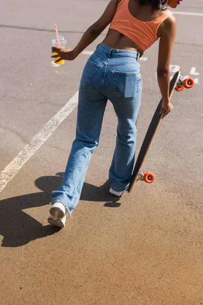 Vista parcial de la mujer afroamericana sosteniendo una taza de plástico con jugo de naranja y longboard afuera - foto de stock