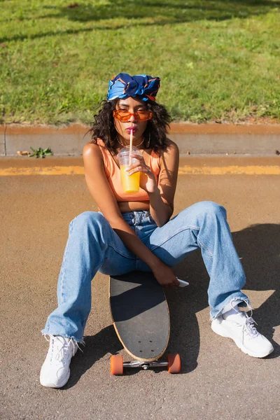 Afroamerikanerin mit Sonnenbrille hält Plastikbecher in der Hand und trinkt Orangensaft, während sie draußen auf dem Longboard sitzt — Stockfoto