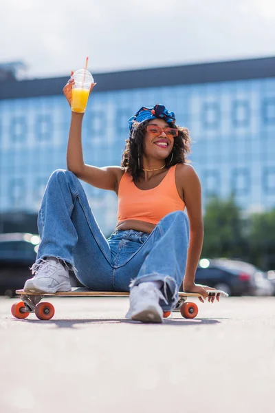 Smiling african american woman in sunglasses sitting on longboard while holding smartphone and plastic cup — Stock Photo