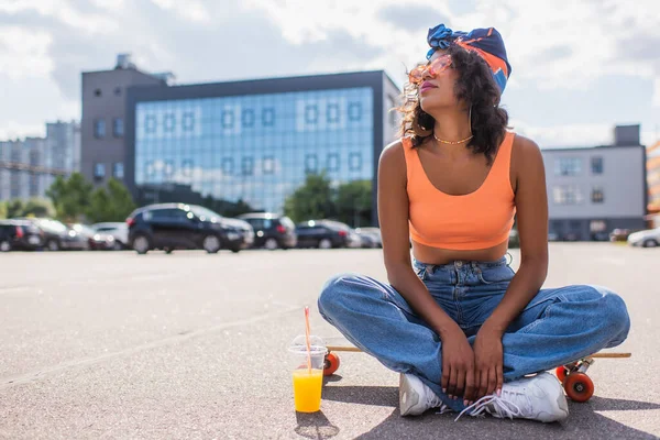 African american woman in sunglasses sitting with crossed legs on longboard near plastic cup with orange juice on asphalt — Stock Photo