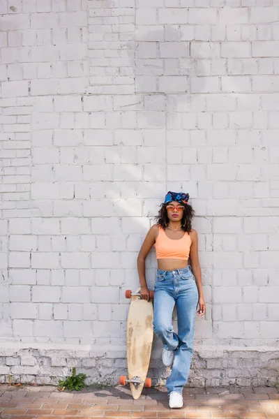 Stylish african american woman in sunglasses and headscarf holding longboard near brick wall — Stock Photo