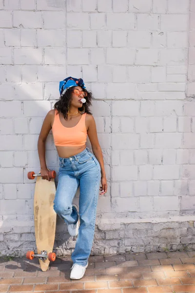 Stylish african american woman in sunglasses and headscarf blowing bubble gum and holding longboard near brick wall — Stock Photo