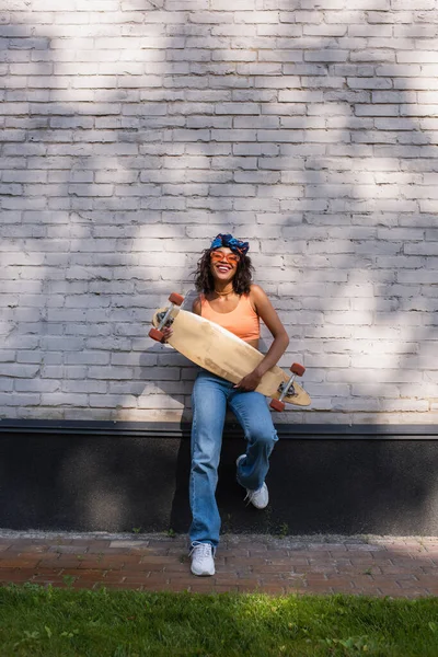 Comprimento total de mulher americana africana feliz em óculos de sol e lenço de cabeça segurando longboard perto da parede de tijolo — Fotografia de Stock