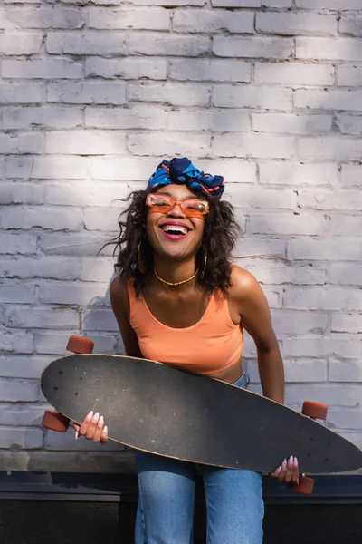 Happy young african american woman in sunglasses and headscarf holding longboard near brick wall — Stock Photo
