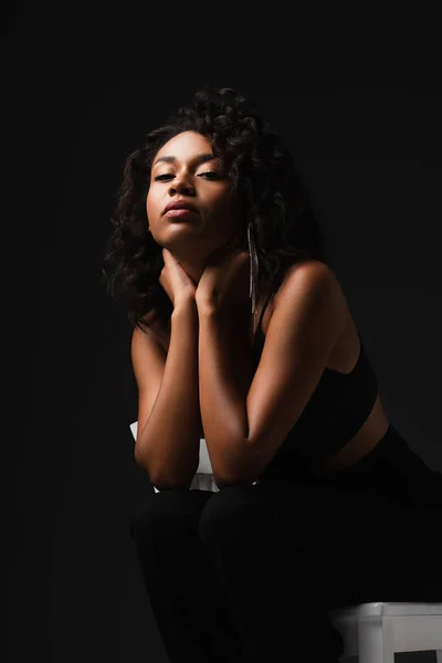 Fashionable african american woman in satin crop top and pants posing on black — Stock Photo
