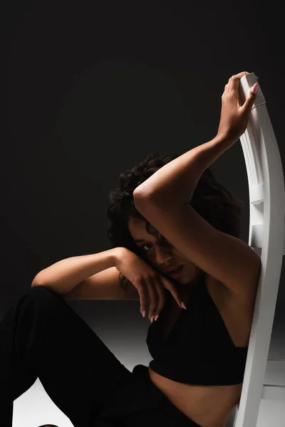 Сексуальна афроамериканка у верхній частині атласу сидить і позує біля білого стільця на чорному — стокове фото