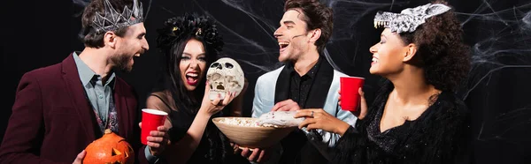 Excited multicultural friends drinking beer and eating popcorn during halloween party on black, banner — Stock Photo