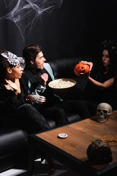 Asiático mujer asustando interracial amigos con jack o linterna cerca bowl de palomitas de maíz en negro - foto de stock