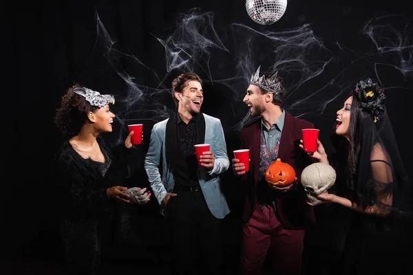 Happy multiethnic friends with plastic cups talking during halloween party on black — Stock Photo