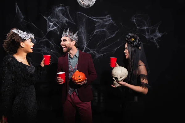 Excited multiethnic friends in halloween costumes holding plastic cups, skull and carved pumpkin while talking on black — Stock Photo