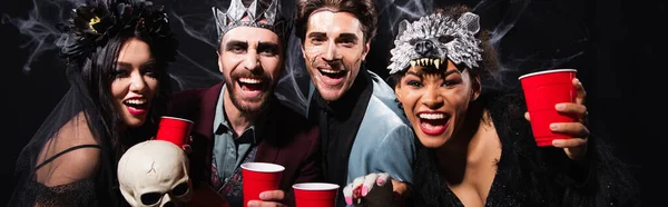 Excited multiethnic friends with plastic cups laughing at camera during halloween party on black, banner — Stock Photo