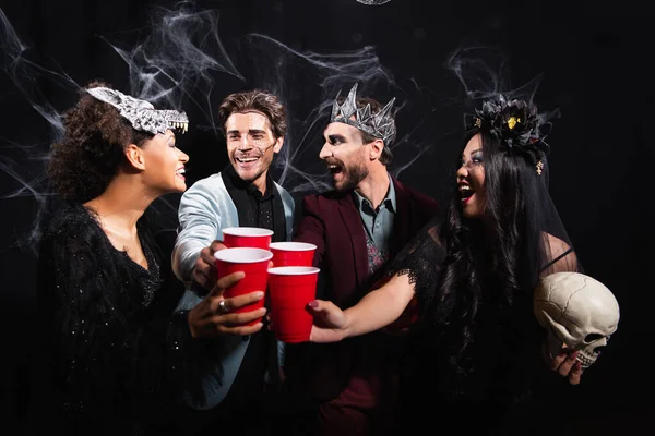 Взволнованные многонациональные друзья в костюмах на Хэллоуин тост с пластиковыми чашками на черном — стоковое фото