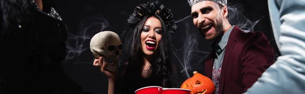 Alegre interracial amigos en vampiros halloween trajes celebración espeluznante cráneo y tallado calabaza en negro, bandera - foto de stock