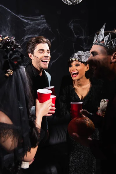 Excited multicultural friends with plastic cup talking during halloween party on black — Stock Photo