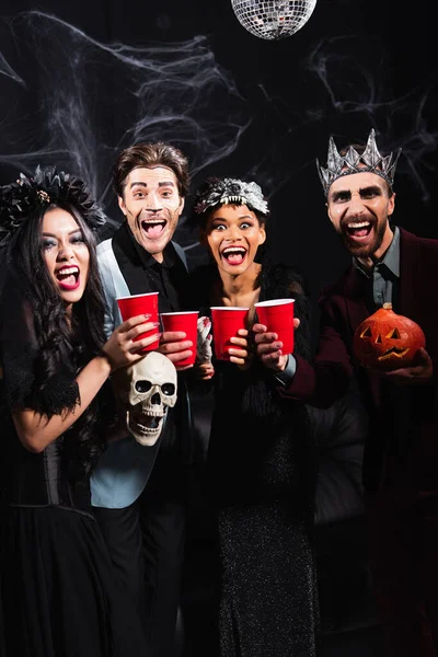 Excited multiethnic people with plastic cups laughing at camera during halloween party on black — Stock Photo