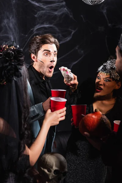 Enojado africano americano mujer en hombre lobo halloween máscara asustando a amigos con espeluznante juguete mano en negro - foto de stock