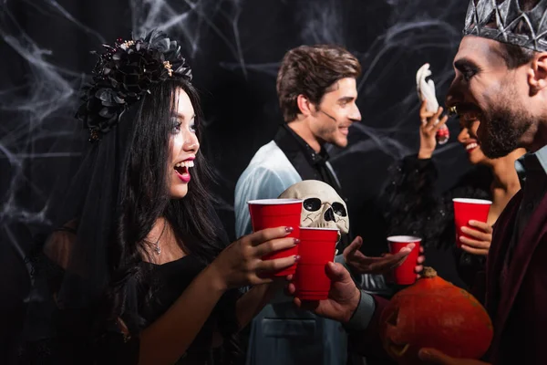 Happy asian woman in black wreath clinking plastic cups with man near blurred friends on black — Stock Photo